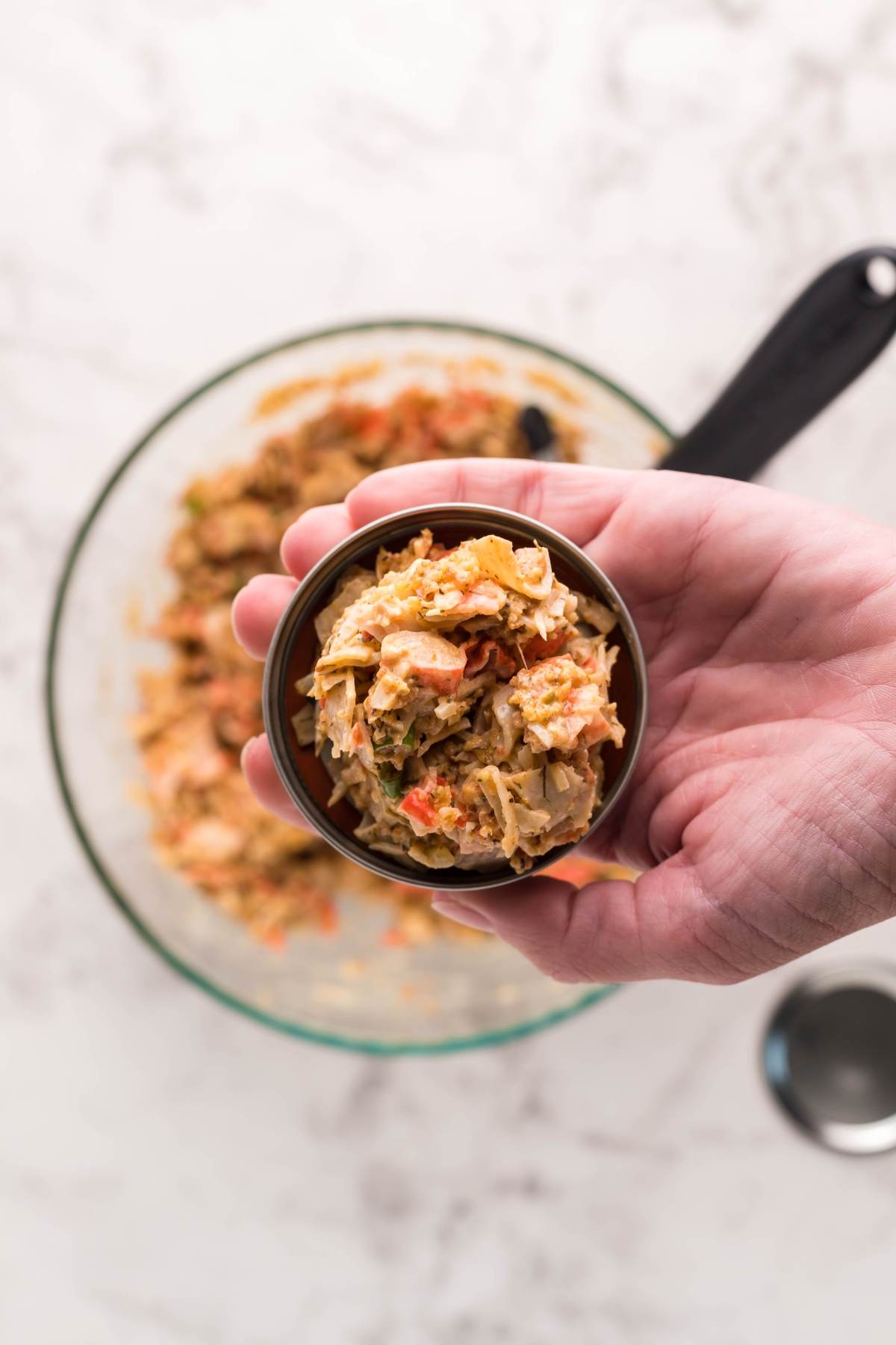 A hand holds a small bowl of vegetable mixture above a larger bowl on a marble surface, preparing ingredients for southwest crab cakes.