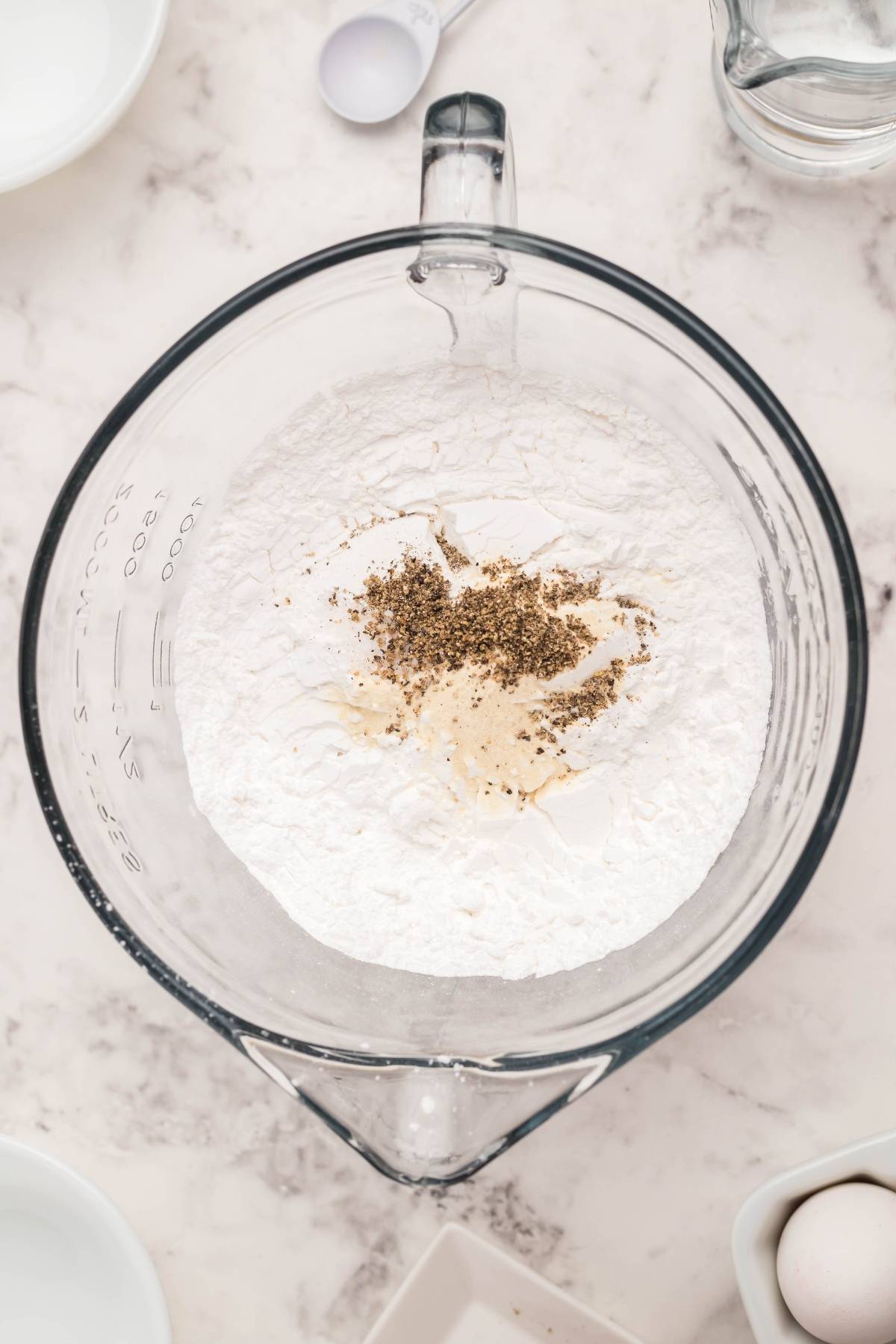 A glass bowl with flour, black pepper, and seasonings on a marble surface, perfect for making homemade egg roll wrappers or trying easy gluten free egg rolls, surrounded by kitchen items.