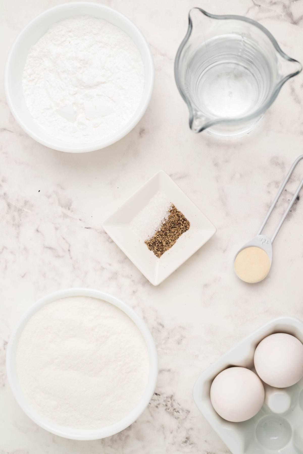 Bowls with flour, water, eggs, yeast, and a dish with salt and pepper on a marble surface—perfect for making homemade egg roll wrappers or trying a tasty air fryer egg rolls recipe.