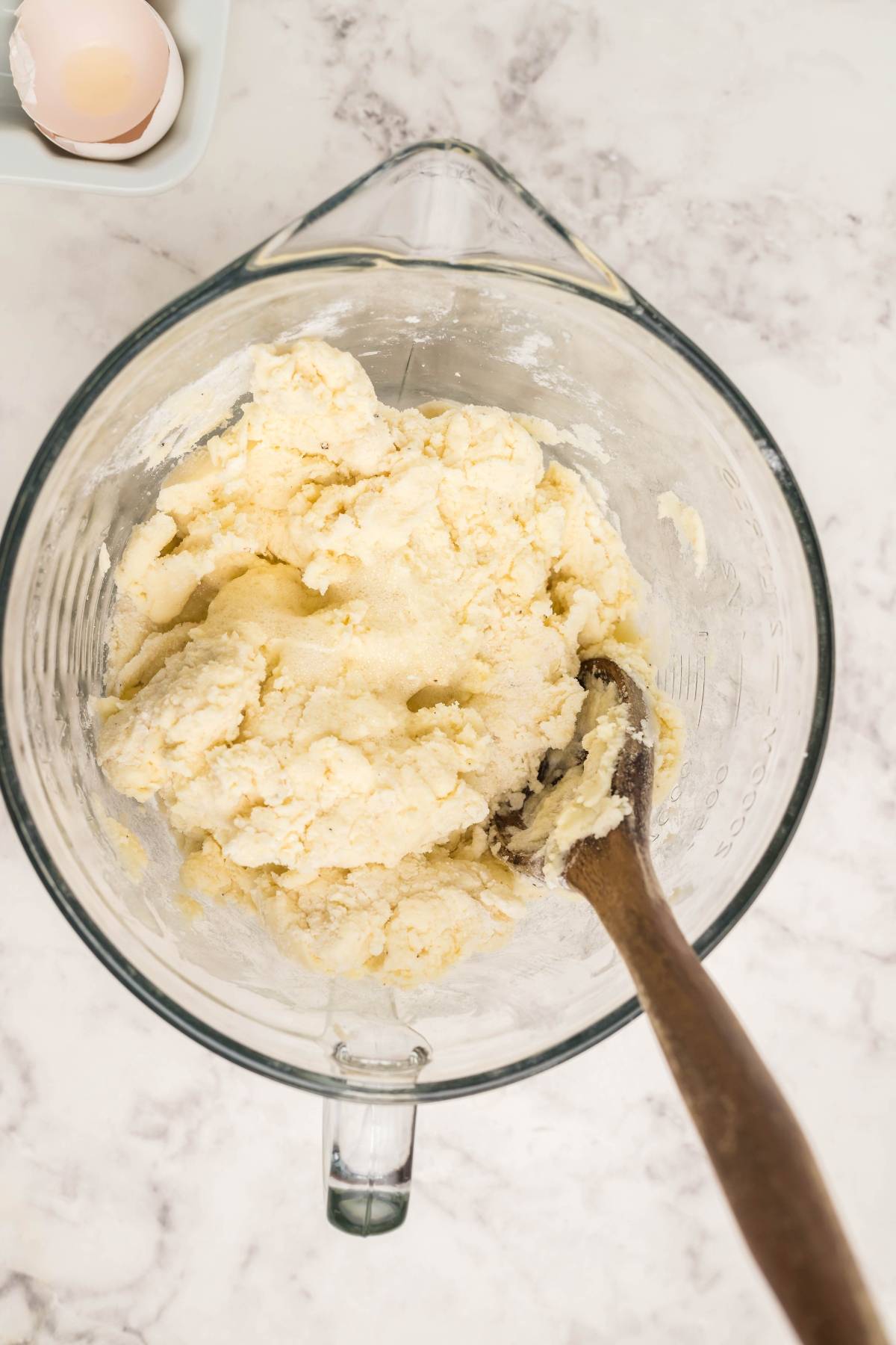 A glass bowl with cookie dough and a wooden spoon sits on a marble countertop beside a cracked egg—perfect for whipping up homemade egg roll wrappers or easy gluten free egg rolls.