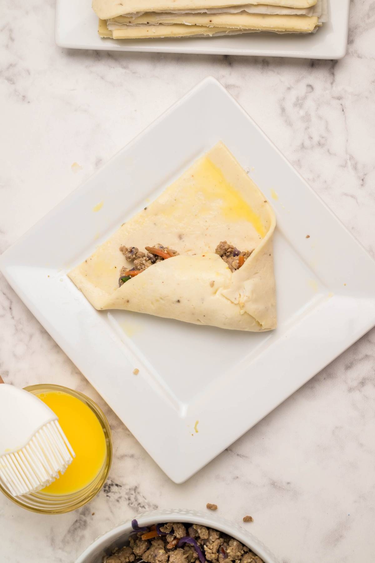 A square plate with a folded pastry filled with meat and vegetables, made using Air Fryer Gluten Free Egg Roll Wrappers, next to a bowl of egg wash and a pastry brush.