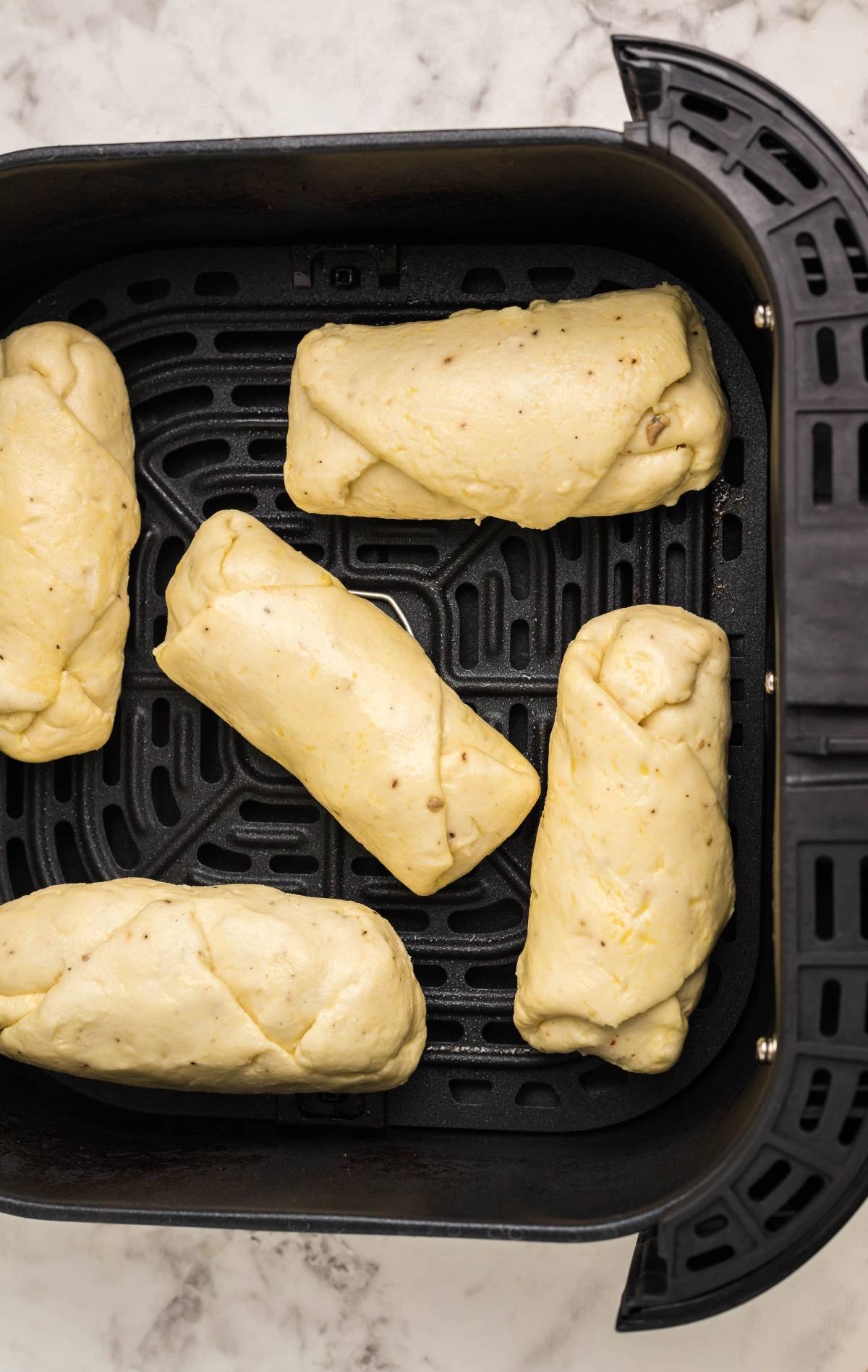 Four uncooked bread rolls placed inside an air fryer basket on a marble countertop—perfect for an easy gluten free egg rolls treat.