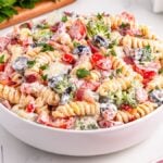 A bowl of creamy bacon ranch pasta salad with rotini, tomatoes, olives, broccoli, and parsley sits on a white table.