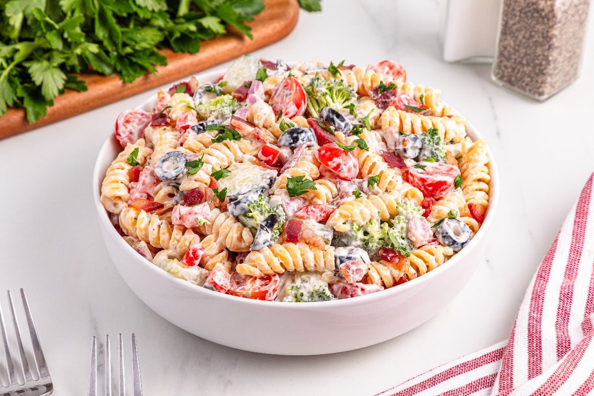 A bowl of creamy bacon ranch pasta salad with rotini, cherry tomatoes, black olives, broccoli, and fresh herbs.