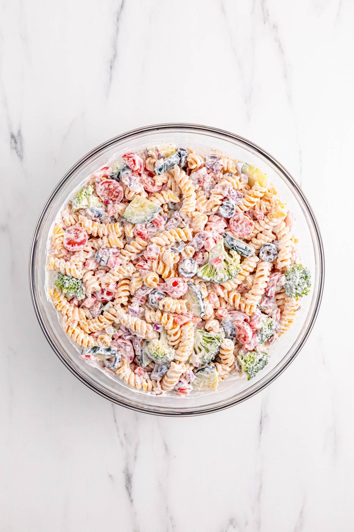 A glass bowl filled with creamy pasta salad, featuring rotini, broccoli, olives, and cherry tomatoes.