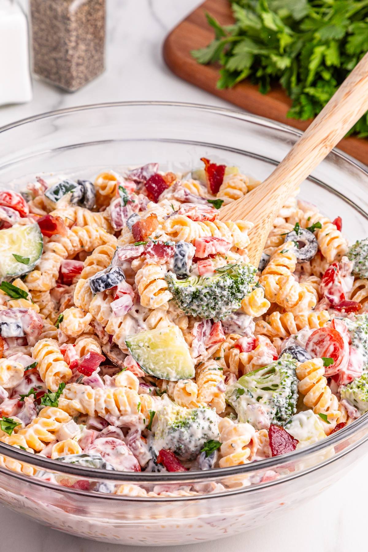 A glass bowl of creamy pasta salad with vegetables, being mixed with a wooden spoon.