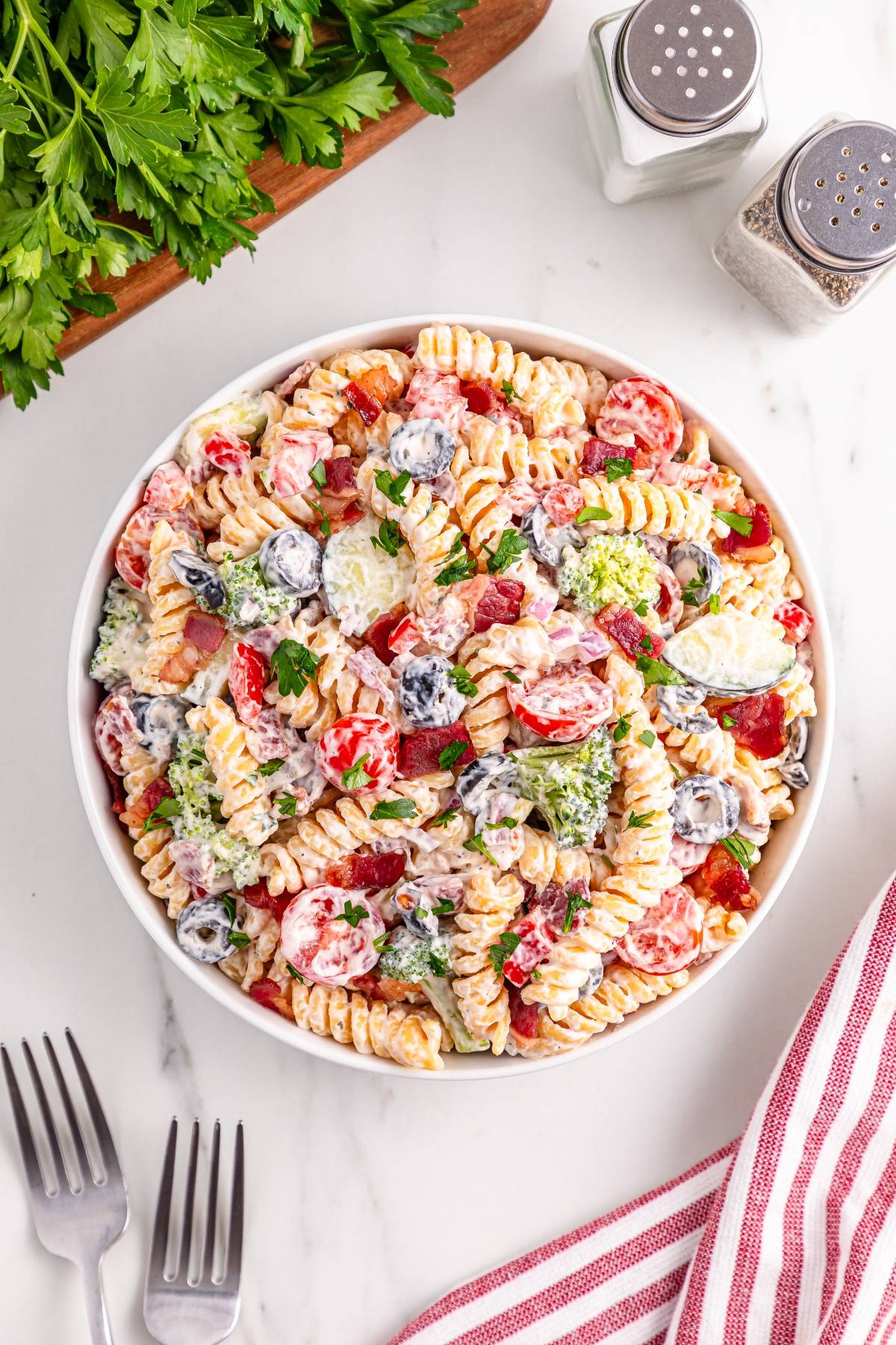 A bowl of creamy pasta salad with vegetables and olives, next to forks, herbs, and salt and pepper shakers.