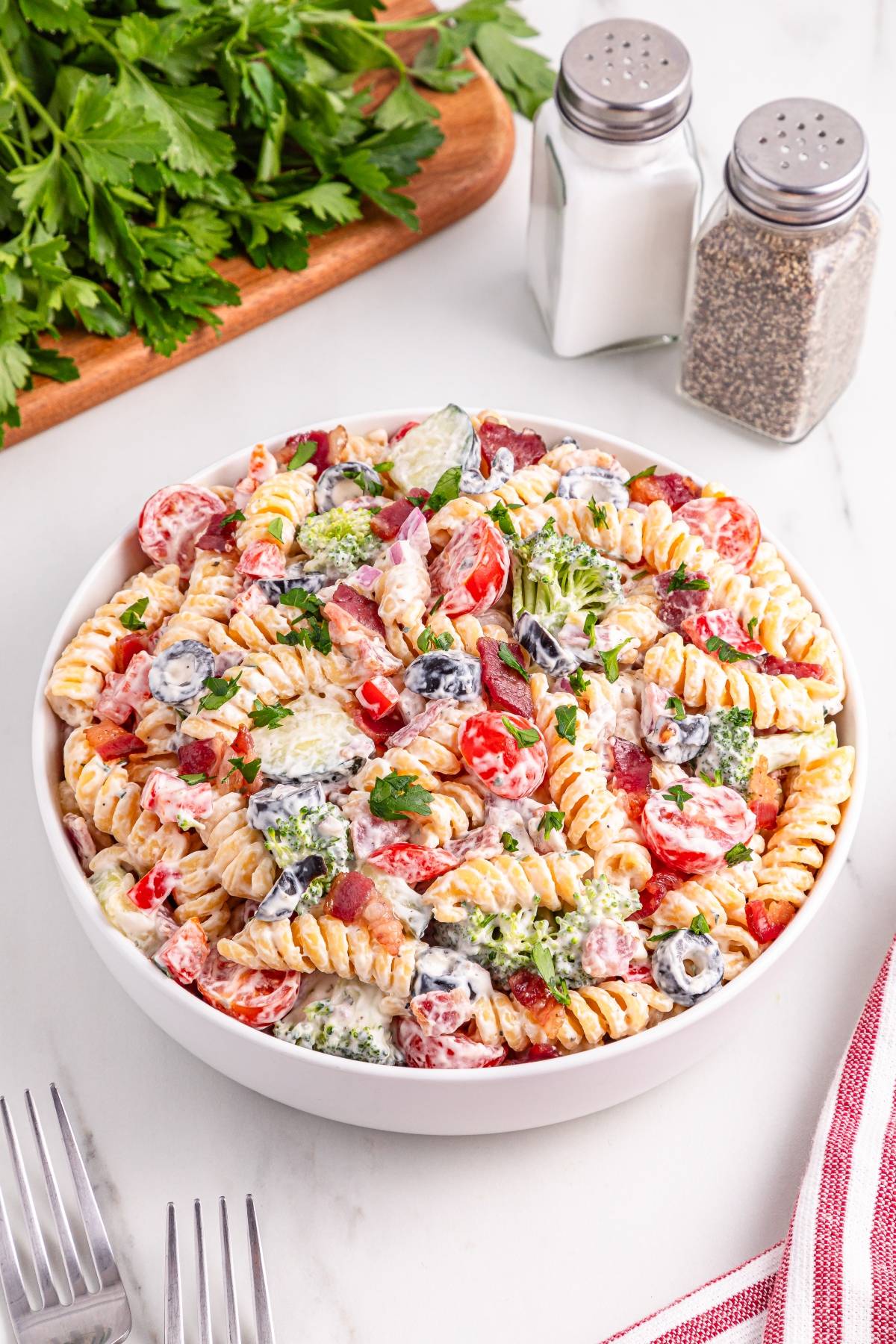 A bowl of creamy pasta salad with tomatoes, olives, and parsley, next to salt, pepper, and fresh herbs.