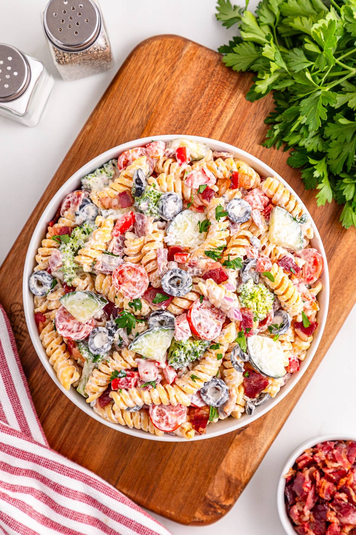 A bowl of creamy rotini pasta salad with tomatoes, olives, cucumbers, and parsley on a wooden board.