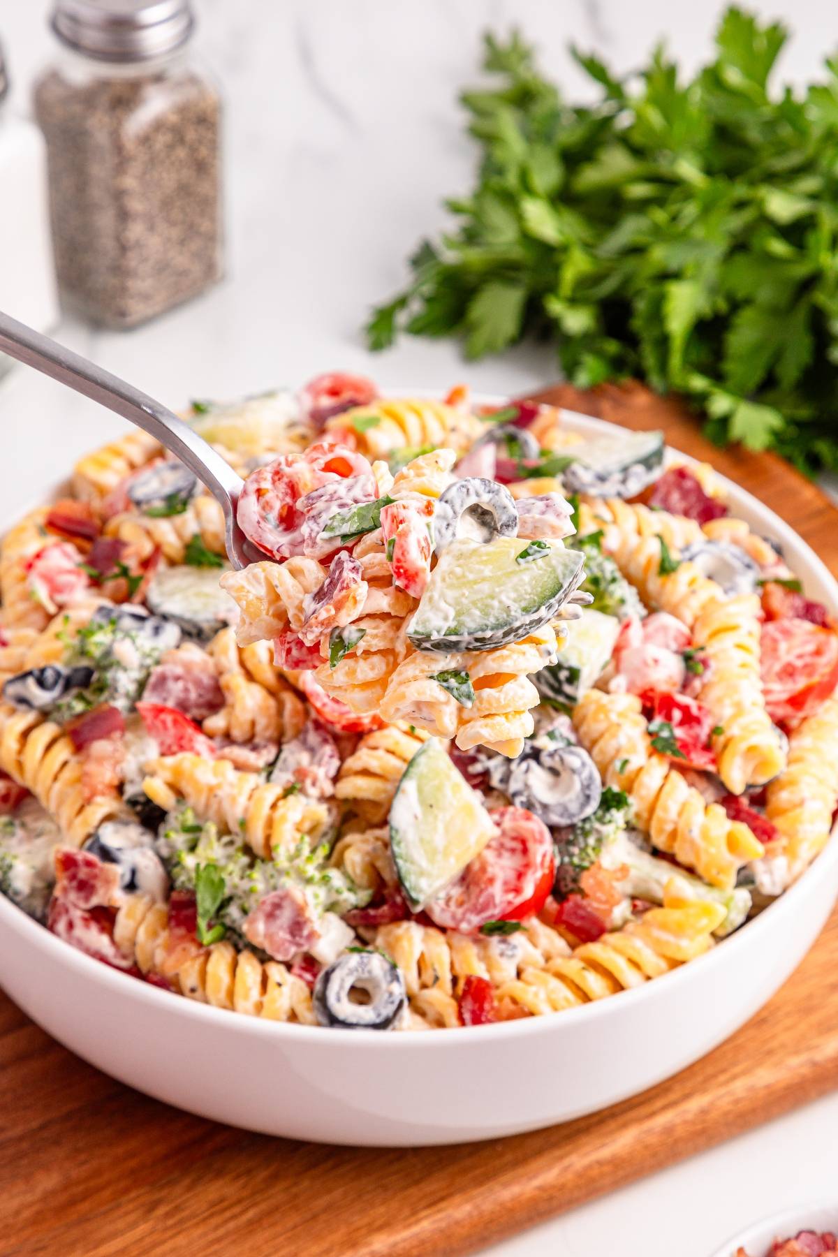 A bowl of creamy pasta salad with rotini, cucumbers, tomatoes, olives, and herbs; a fork lifts a bite.