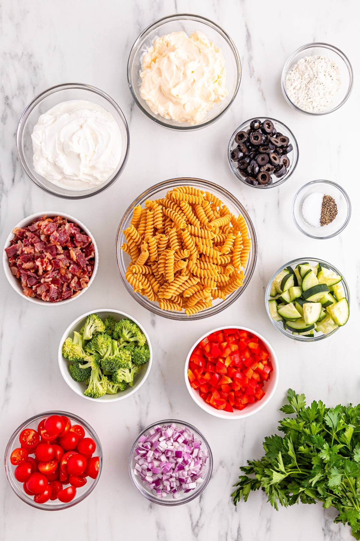 Bowls of pasta, chopped vegetables, bacon, olives, shredded cheese, spices, and parsley on a marble surface.