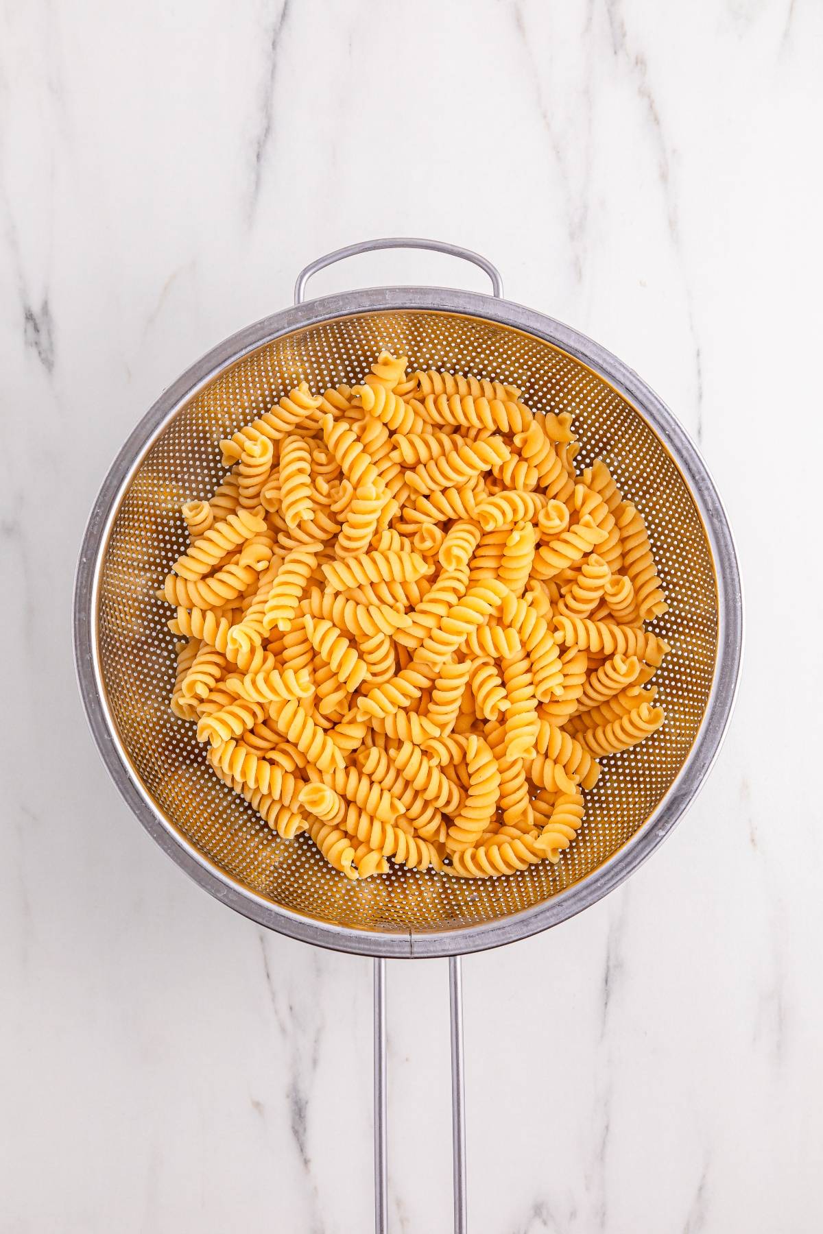 Cooked rotini pasta draining in a metal colander on a white marble surface.