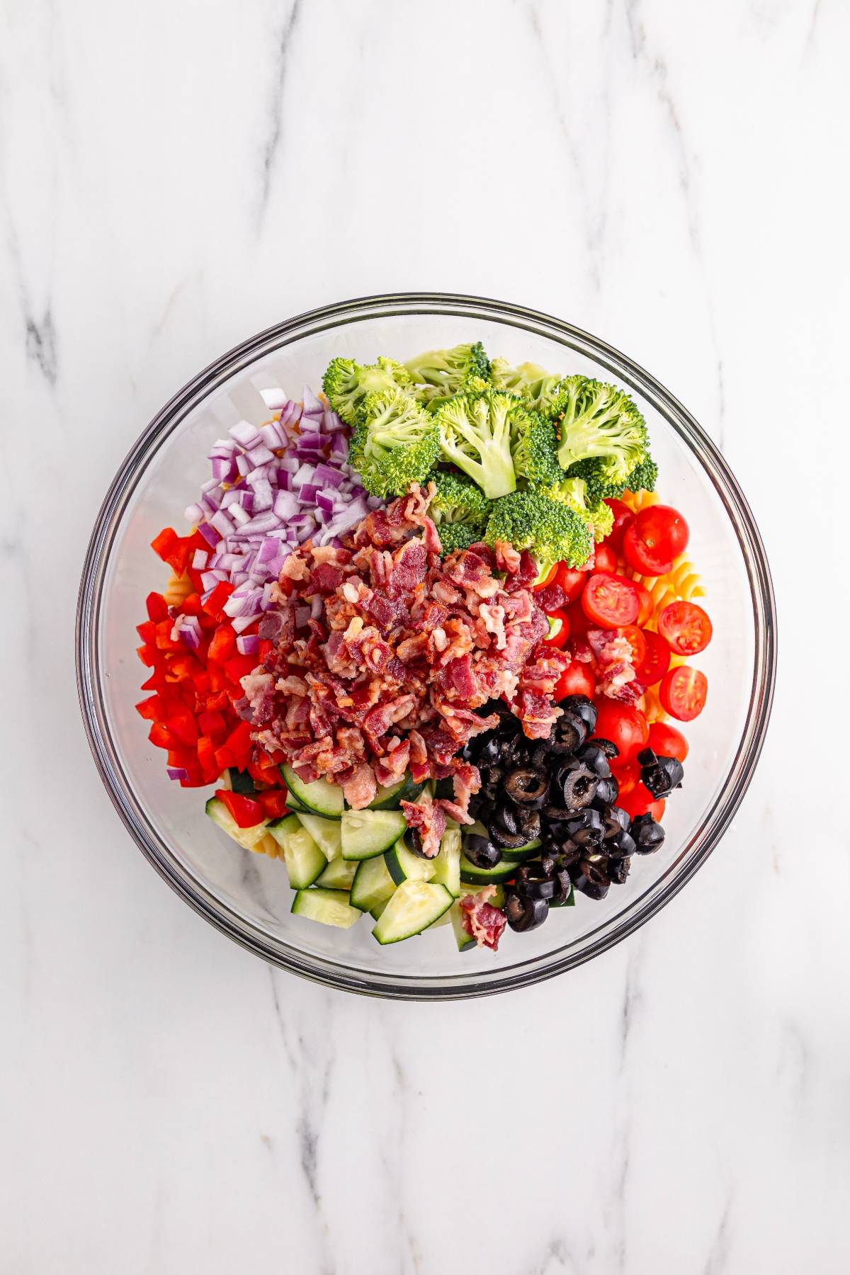 Glass bowl with chopped bacon, broccoli, tomatoes, red onion, red pepper, cucumber, and black olives.