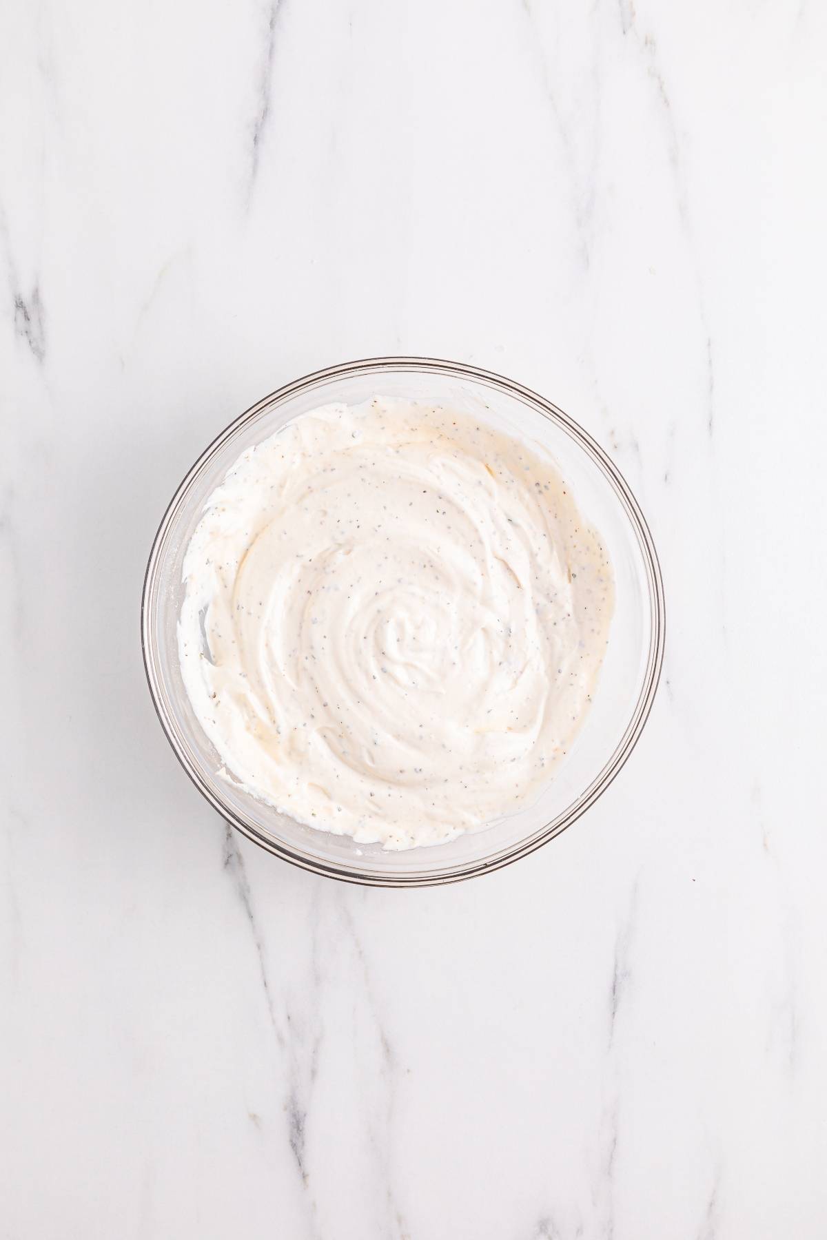 A glass bowl filled with creamy white sauce sits on a white marble surface.