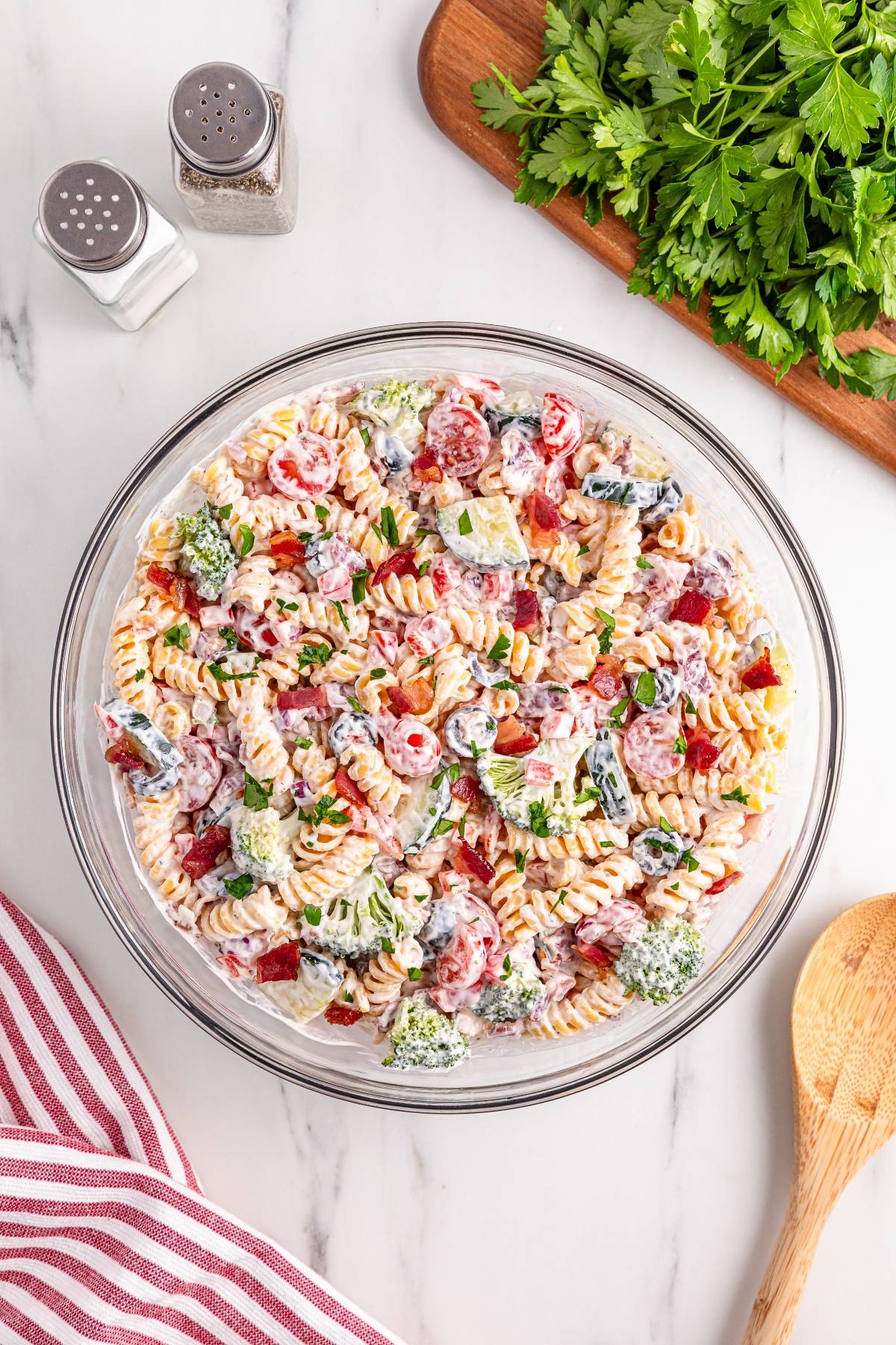 A bowl of creamy pasta salad with vegetables, next to fresh parsley and salt and pepper shakers.