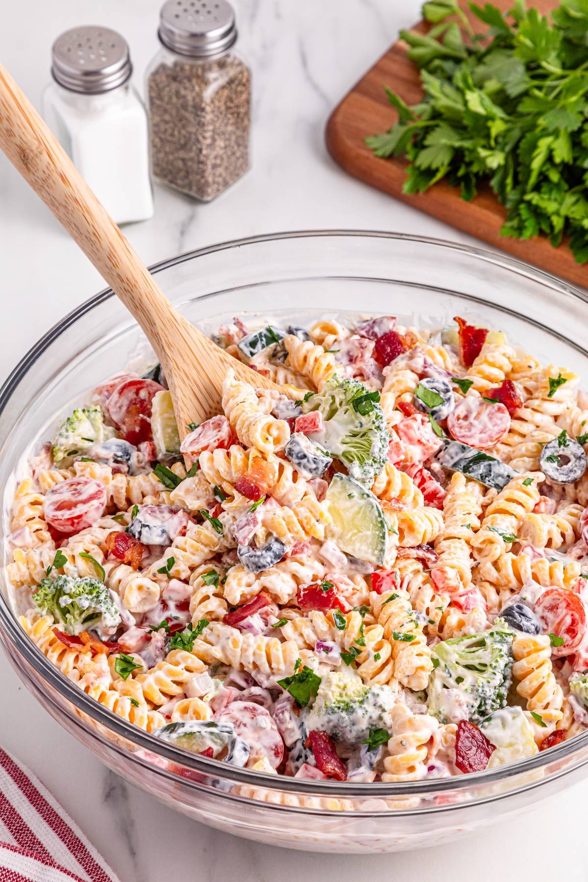 A bowl of creamy pasta salad with rotini, tomatoes, cucumber, olives, and broccoli, mixed with a wooden spoon.