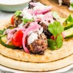 Two pita breads topped with beef kofta, cherry tomatoes, cucumber, pickled onions, yogurt sauce, and cilantro.