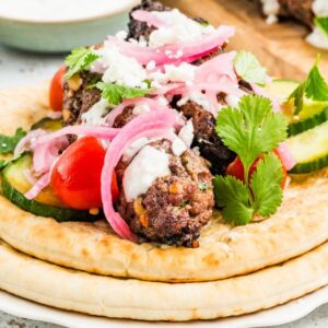 Two pita breads topped with beef kofta, cherry tomatoes, cucumber, pickled onions, yogurt sauce, and cilantro.