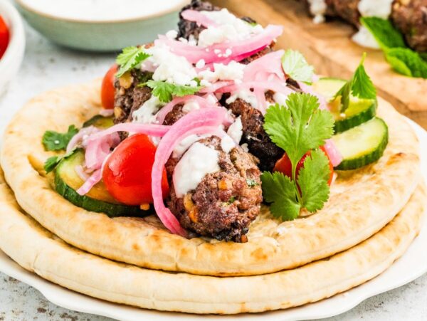 Two pita breads topped with beef kofta, cherry tomatoes, cucumber, pickled onions, yogurt sauce, and cilantro.