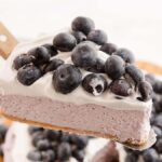 A slice of no bake blueberry cheesecake with whipped cream and fresh blueberries on top is being served.