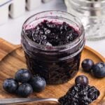 A jar of blueberry jam sits on a wooden board, surrounded by fresh blueberries and a spoonful of rich, homemade blueberry jam.
