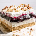 A slice of Blueberry Lush dessert with whipped cream and chopped nuts, lifted on a spatula.