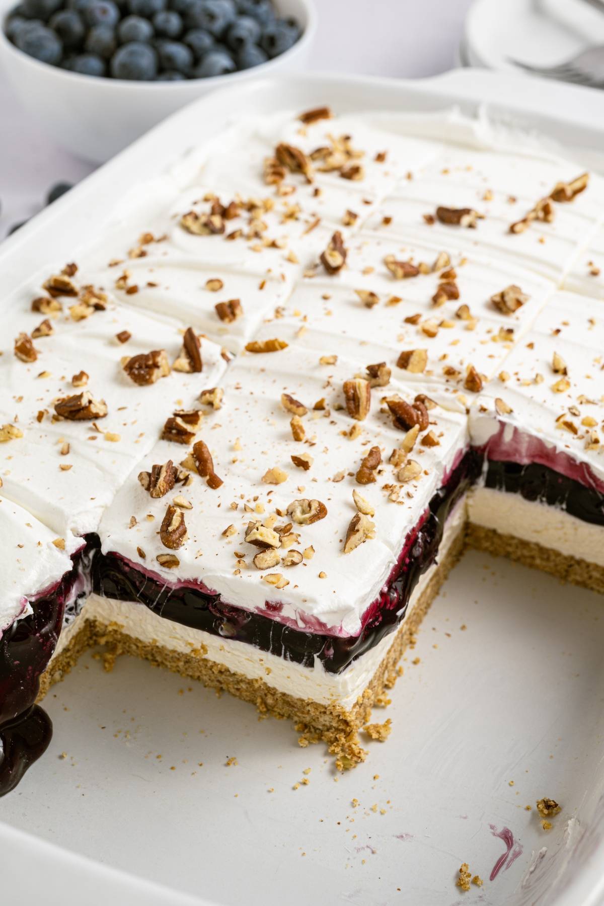 A layered dessert with a graham crust, creamy filling, blueberry topping, whipped cream, and chopped pecans.
