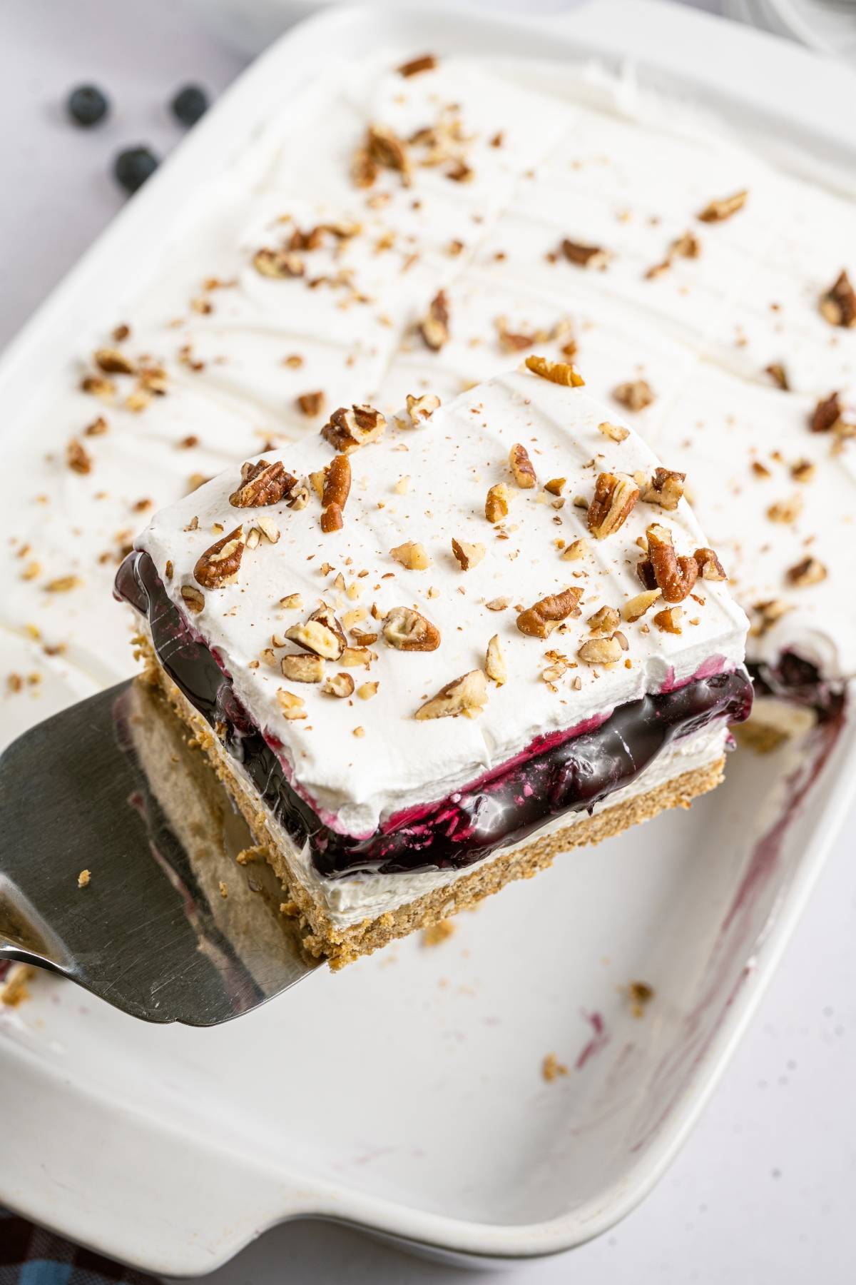 A spatula lifts a slice of layered dessert with whipped cream, chocolate, berry filling, and chopped nuts.