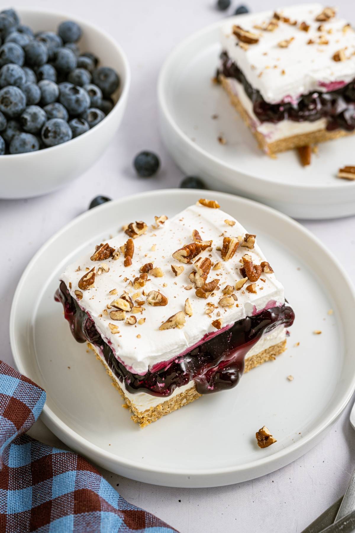 A dessert bar with whipped topping, blueberry filling, and chopped pecans on a plate near a bowl of fresh blueberries.