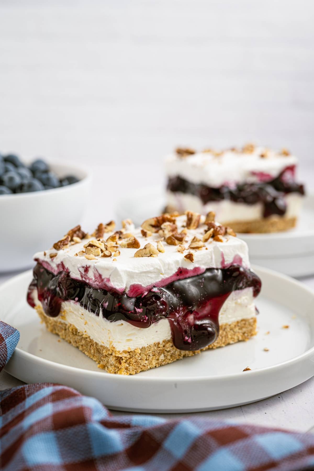 A slice of layered blueberry dessert with whipped topping and pecans on a white plate, with a bowl of blueberries nearby.