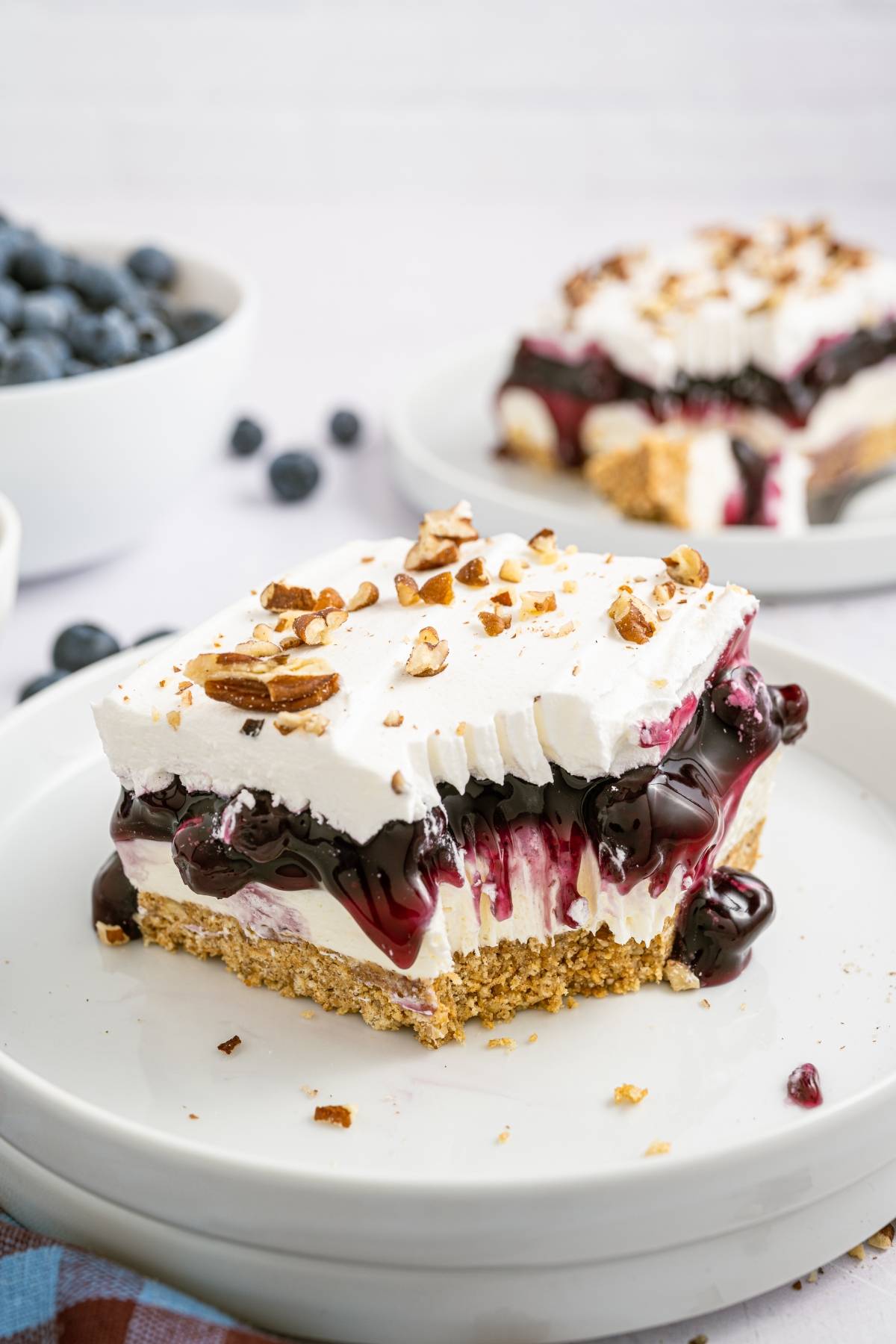 A square slice of blueberry dessert with whipped topping and chopped nuts on a plate, with blueberries in the background.