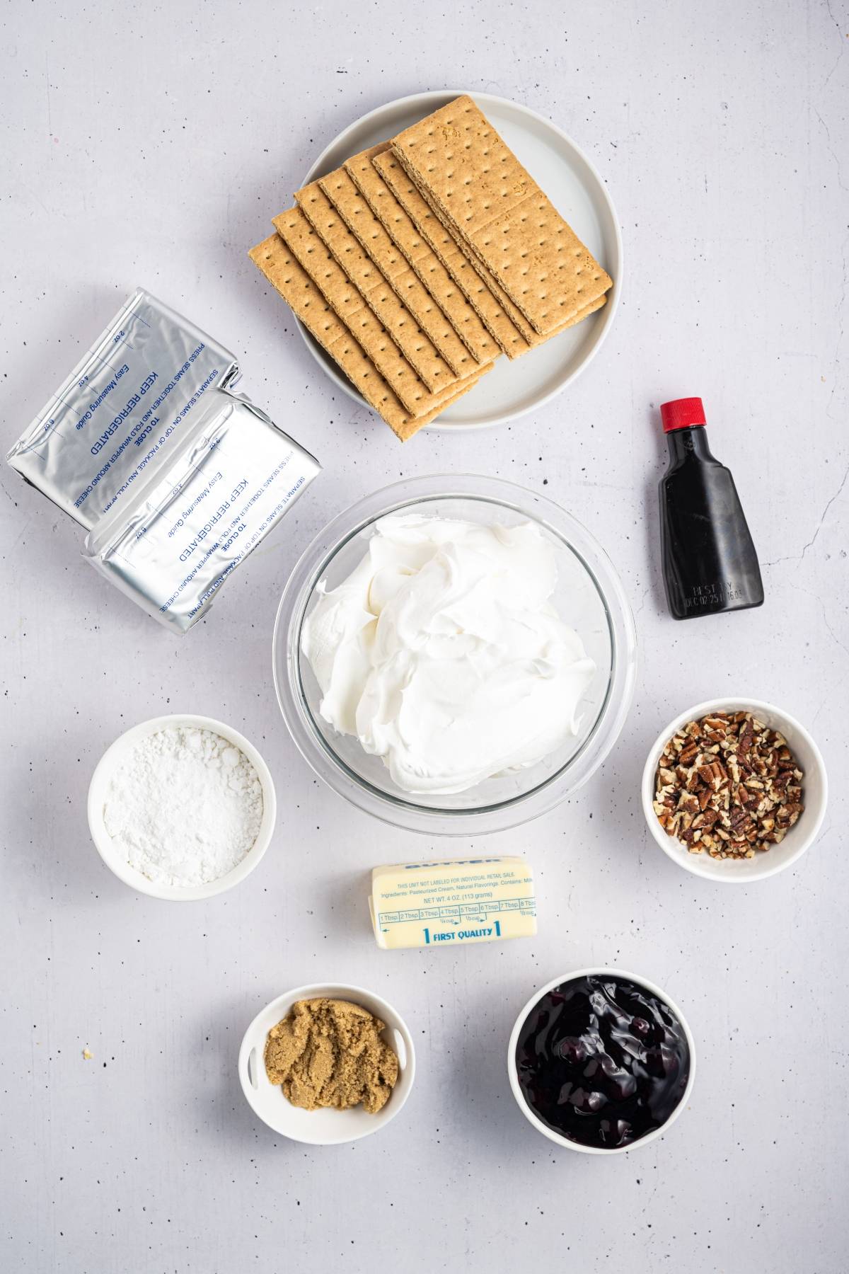 Ingredients for a dessert, including graham crackers, cream cheese, whipped topping, and various bowls of toppings.