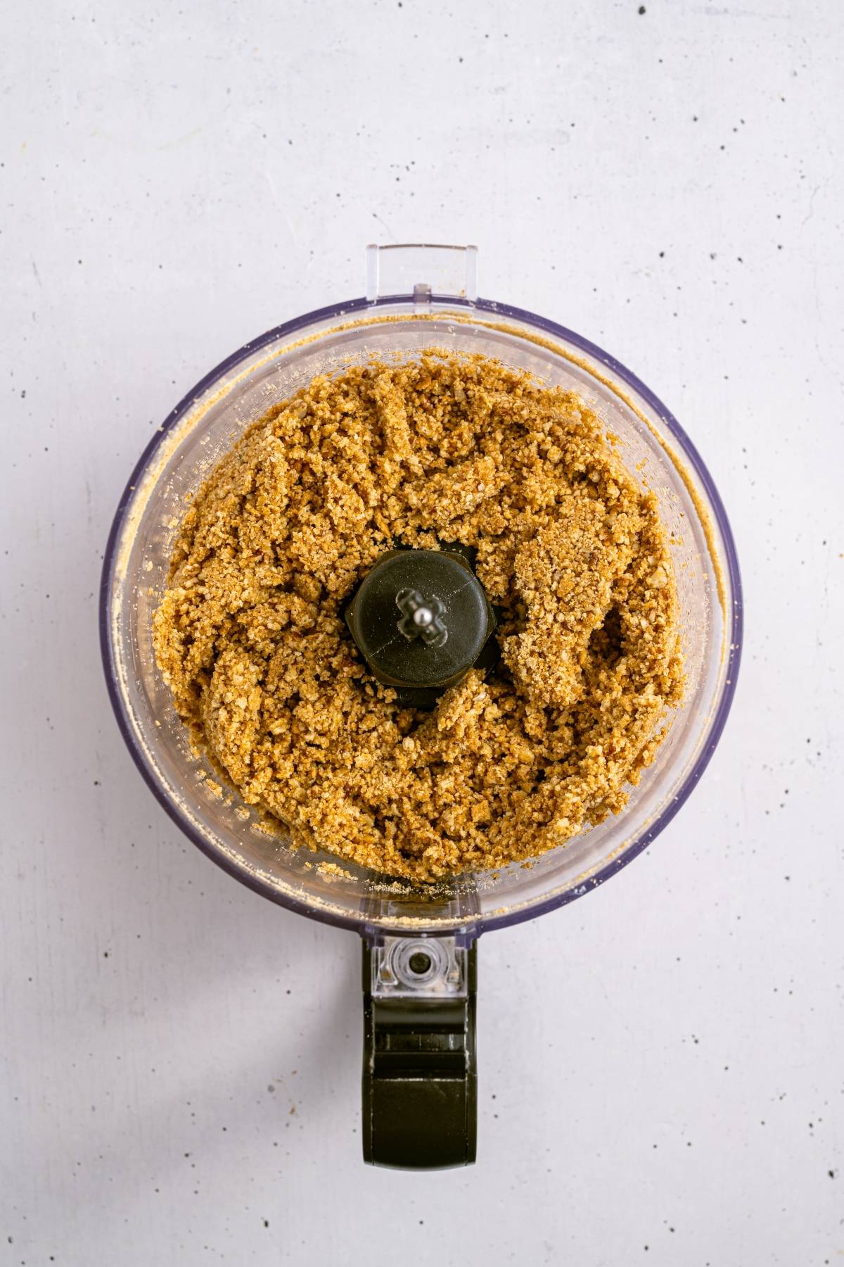 A food processor with finely ground nut mixture inside, viewed from above on a light surface.