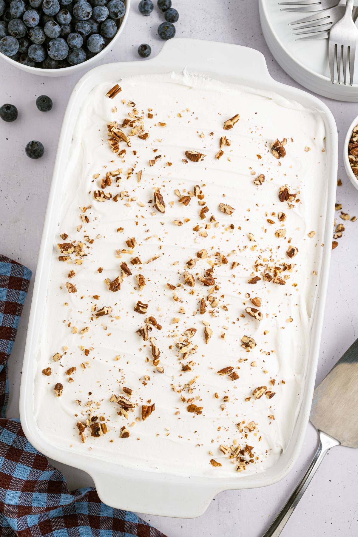 White frosted dessert in a casserole dish topped with chopped nuts, surrounded by blueberries and plates.