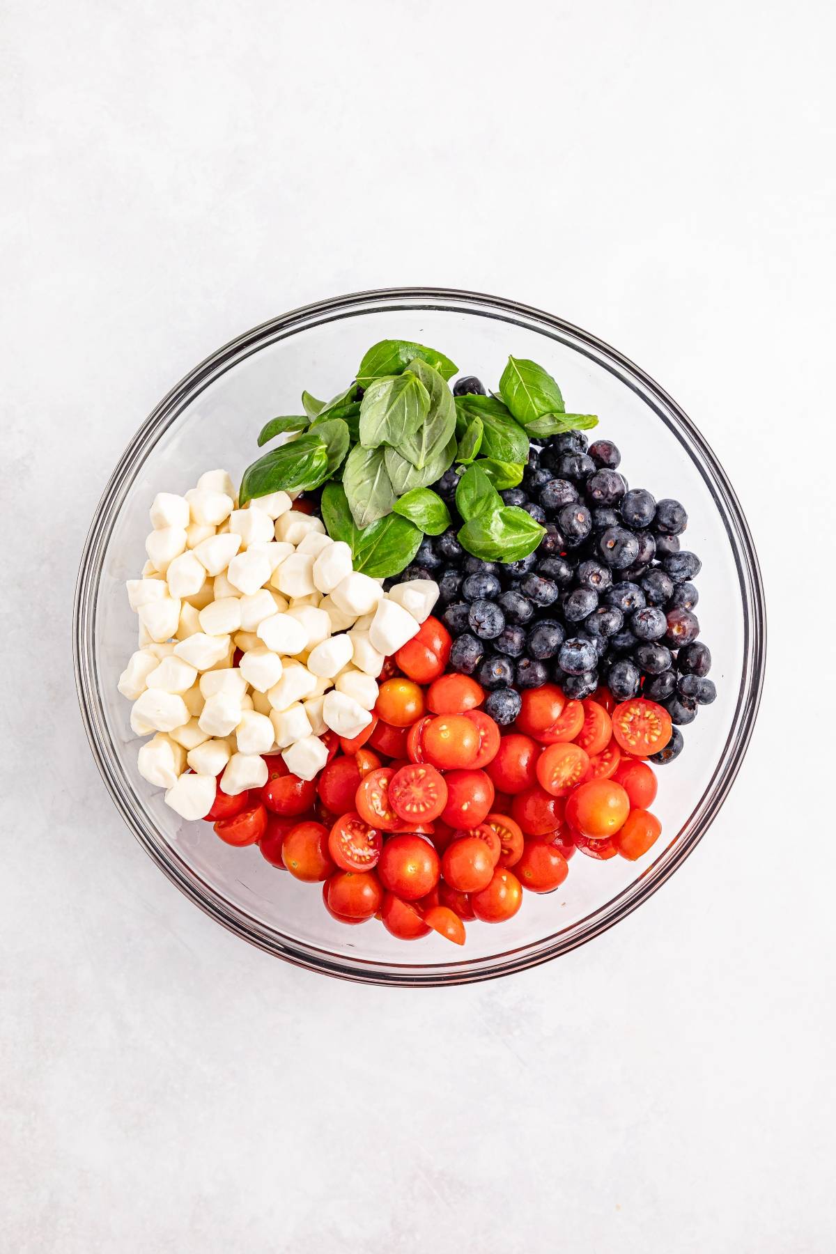 A bowl with mozzarella balls, cherry tomatoes, blueberries, and fresh basil on a white surface.