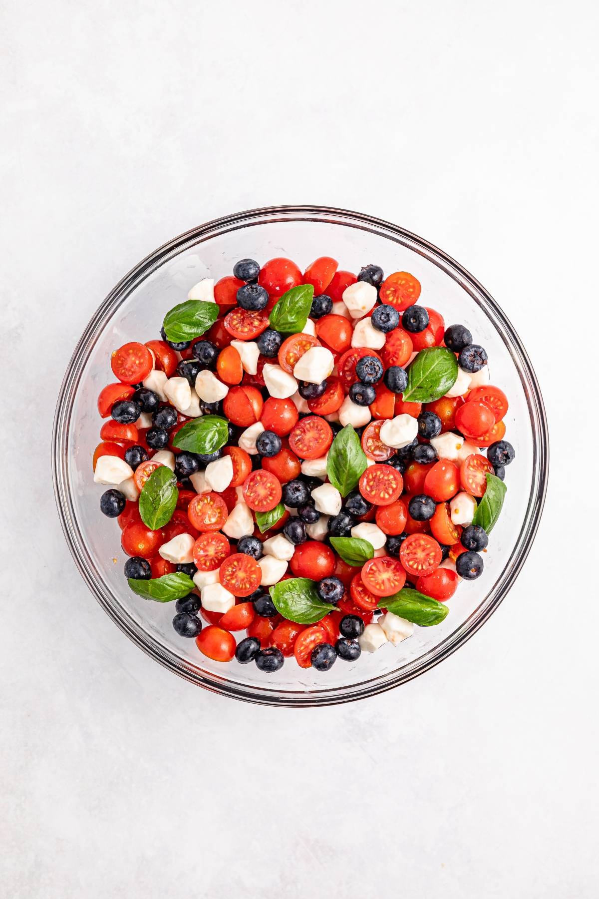 Glass bowl with cherry tomatoes, blueberries, mozzarella balls, and basil leaves on a white background.