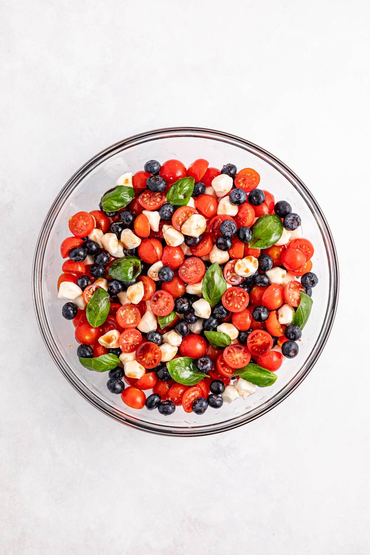 A glass bowl filled with cherry tomatoes, blueberries, mozzarella balls, and fresh basil leaves.