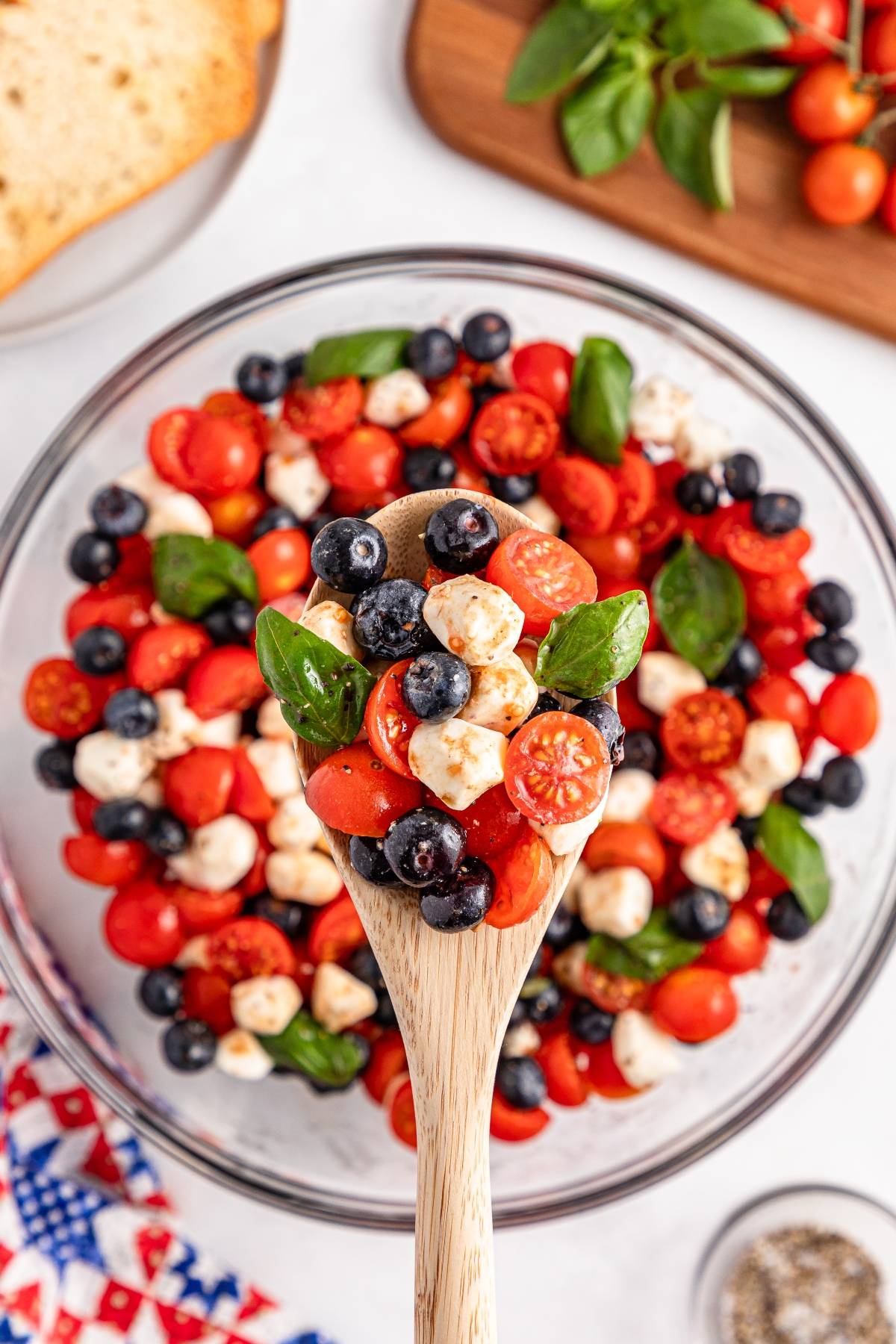 A wooden spoon holds a mix of cherry tomatoes, blueberries, mozzarella balls, and basil over a salad bowl.