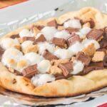 A campfire s'mores pizza featuring flatbread topped with chocolate, marshmallows, and graham cracker pieces on foil.