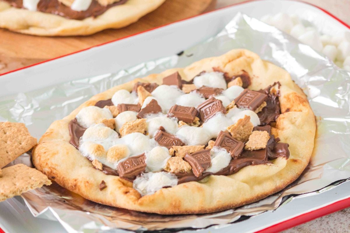 Naan bread topped with melted chocolate, marshmallows, and graham cracker pieces on a foil-lined tray—just like a campfire s'mores pizza.