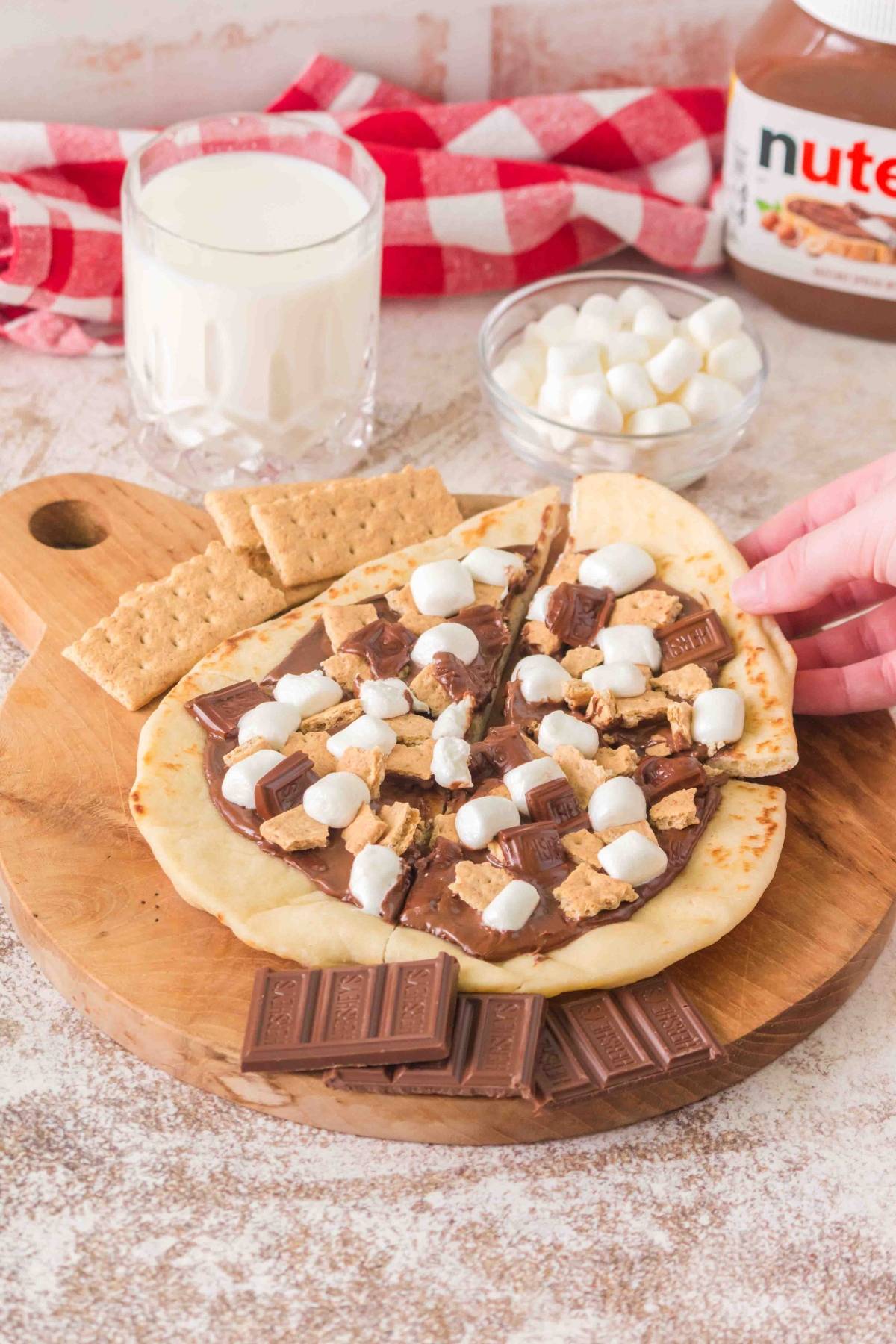 Flatbread topped with chocolate, graham crackers, and marshmallows sits on a board beside milk and Nutella.