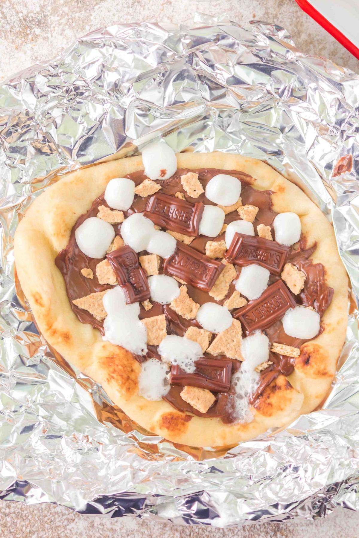 Flatbread topped with melted chocolate, marshmallows, graham cracker pieces on foil.