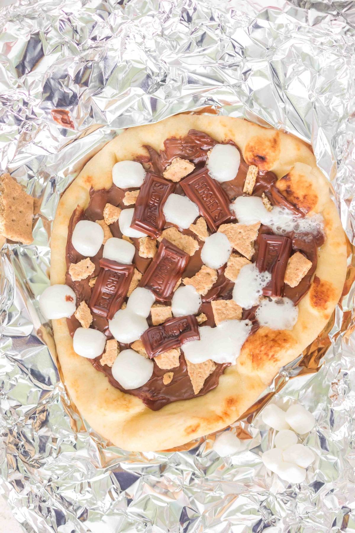 Flatbread topped with chocolate, marshmallows, and graham cracker pieces on foil.