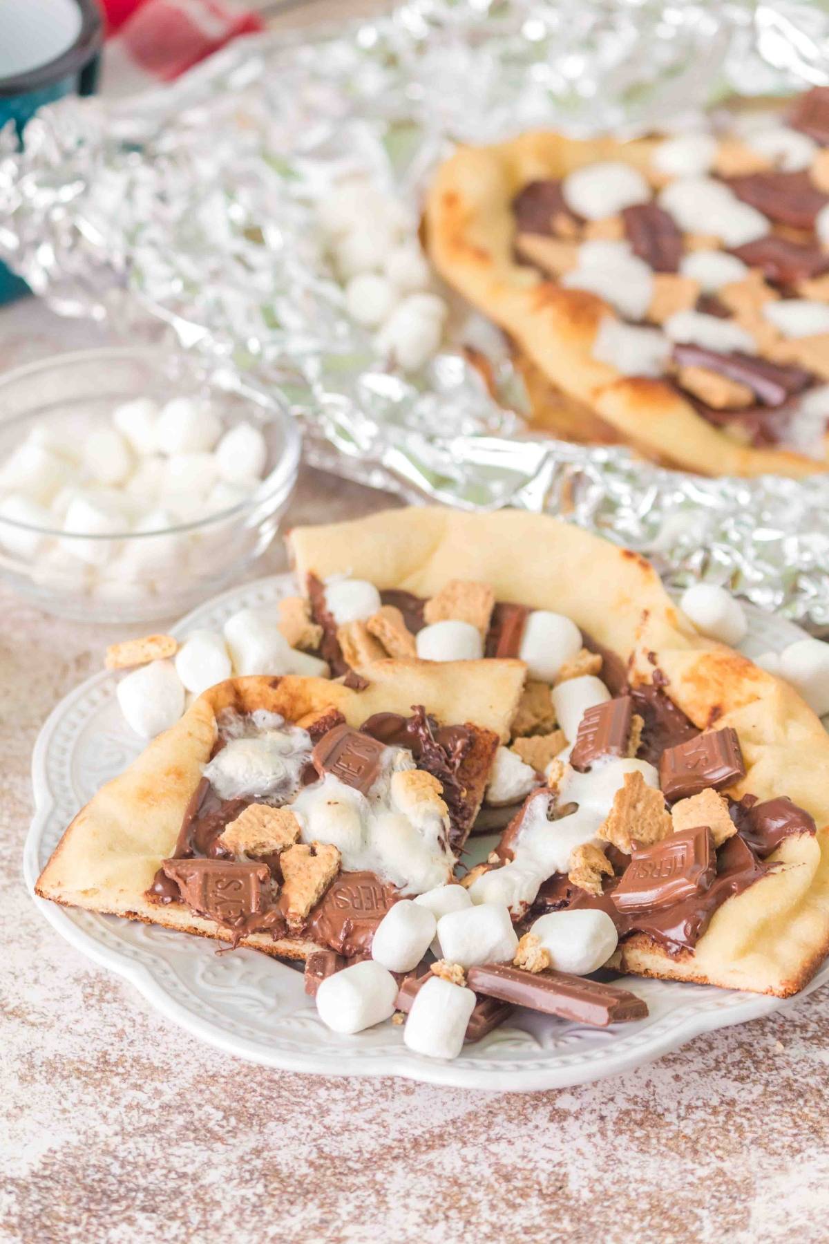 A slice of s'mores pizza with chocolate, marshmallows, and graham crackers on a plate.