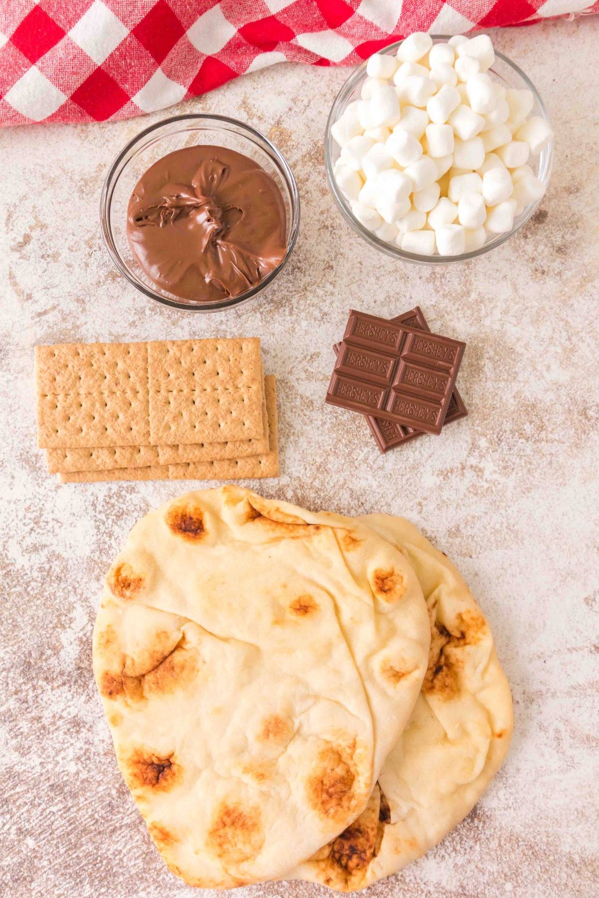 Flatbread, graham crackers, chocolate, Nutella, and mini marshmallows on a light surface with a red checkered cloth.