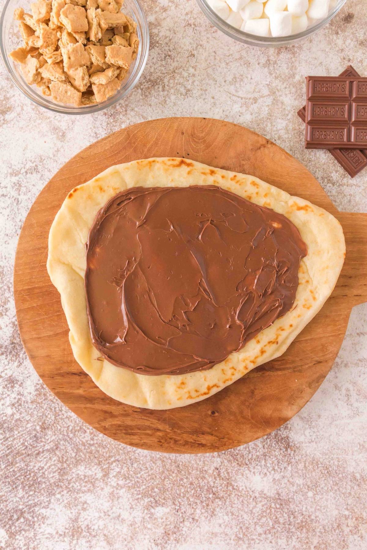Flatbread with chocolate spread on a wooden board, next to graham crackers, marshmallows, and chocolate pieces.