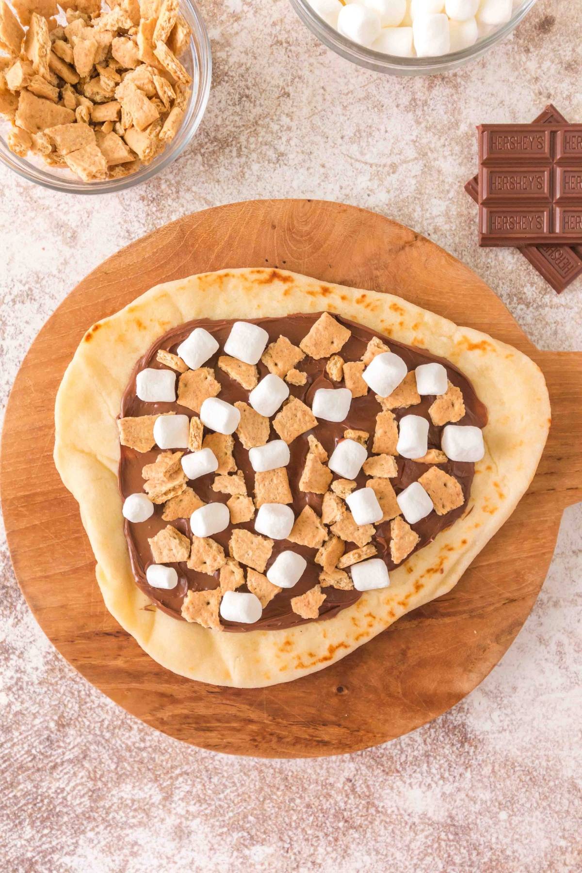 Naan bread topped with chocolate spread, marshmallows, and graham cracker pieces on a wooden board.