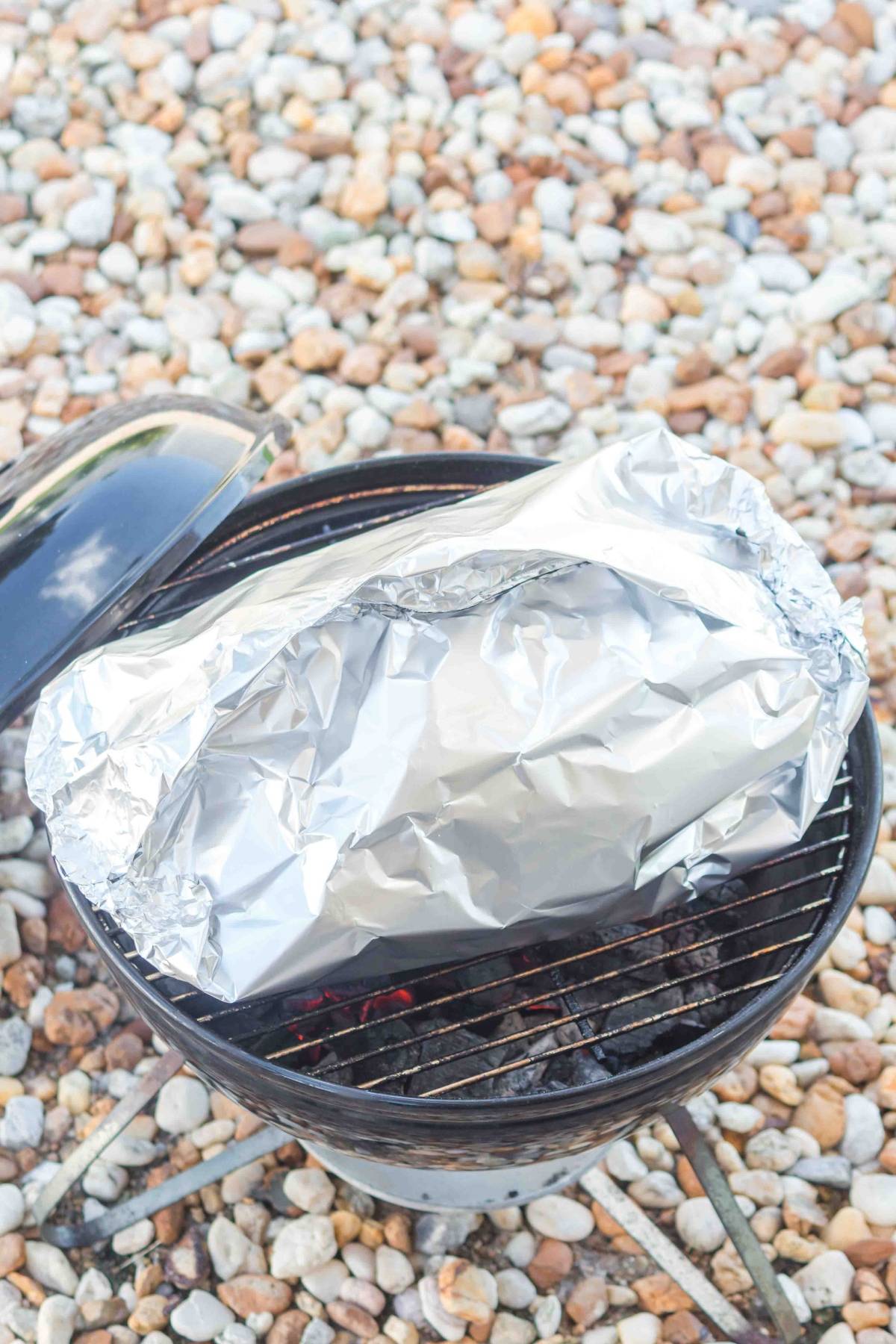 A foil-wrapped package sits on a small charcoal grill over hot coals, with rocks in the background.
