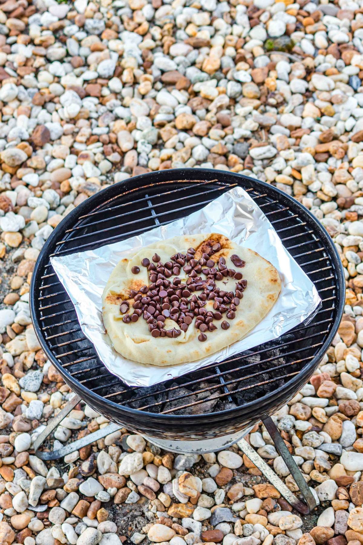 Flatbread topped with chocolate chips on foil, grilling on a small barbecue over rocks.