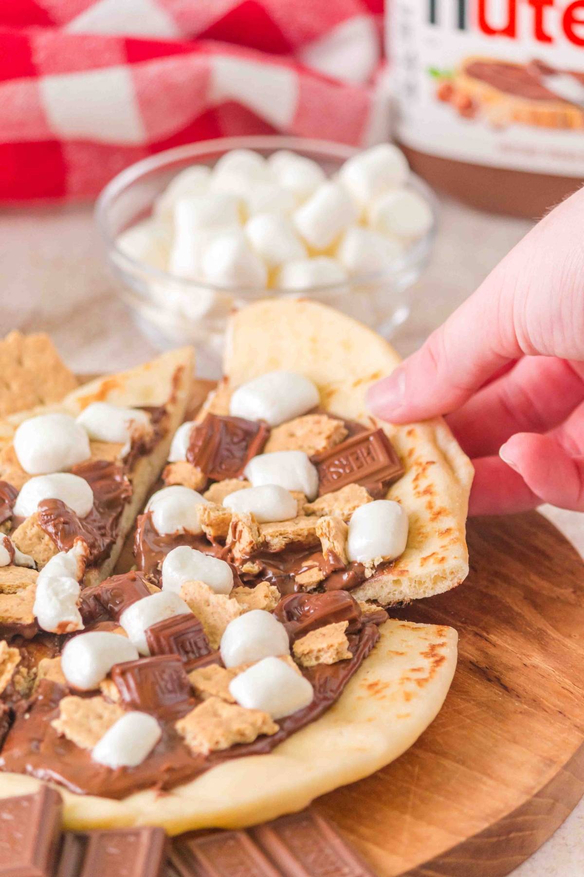 A hand picks up a slice of s'mores pizza topped with chocolate, marshmallows, and graham crackers.